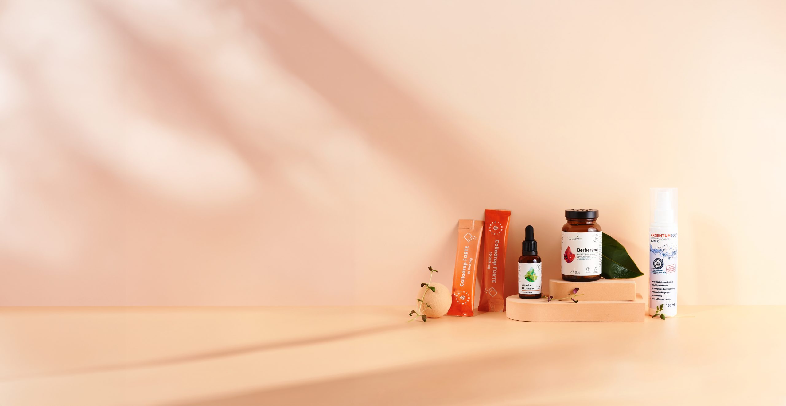Produkty Aura Herbals na jasnopomarańczowym tle: 2 saszetki Colladrop Forte, Witamina B complex w krpolach, Berberyna w kapsułkach i Srebro koloidalne aerozolu