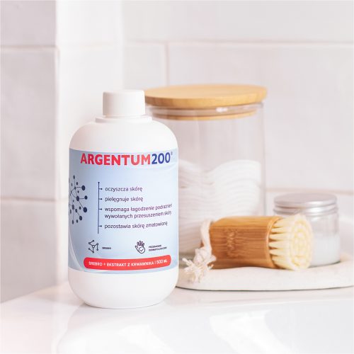 Butelka argentum-200-srebro-krwawnik-500ml stoi obok płatków kosmetycznych, słoika, puszki i drewnianej szczotki na białej powierzchni łazienki.