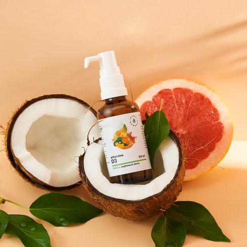 Butelka Witamina-D3-2000-IU-Vegan-krople-50-ml_1500x1500 znajduje się wewnątrz przepołowionego orzecha kokosowego, otoczonego zielonymi liśćmi i plasterkami grejpfruta, a wszystko to na beżowej powierzchni.